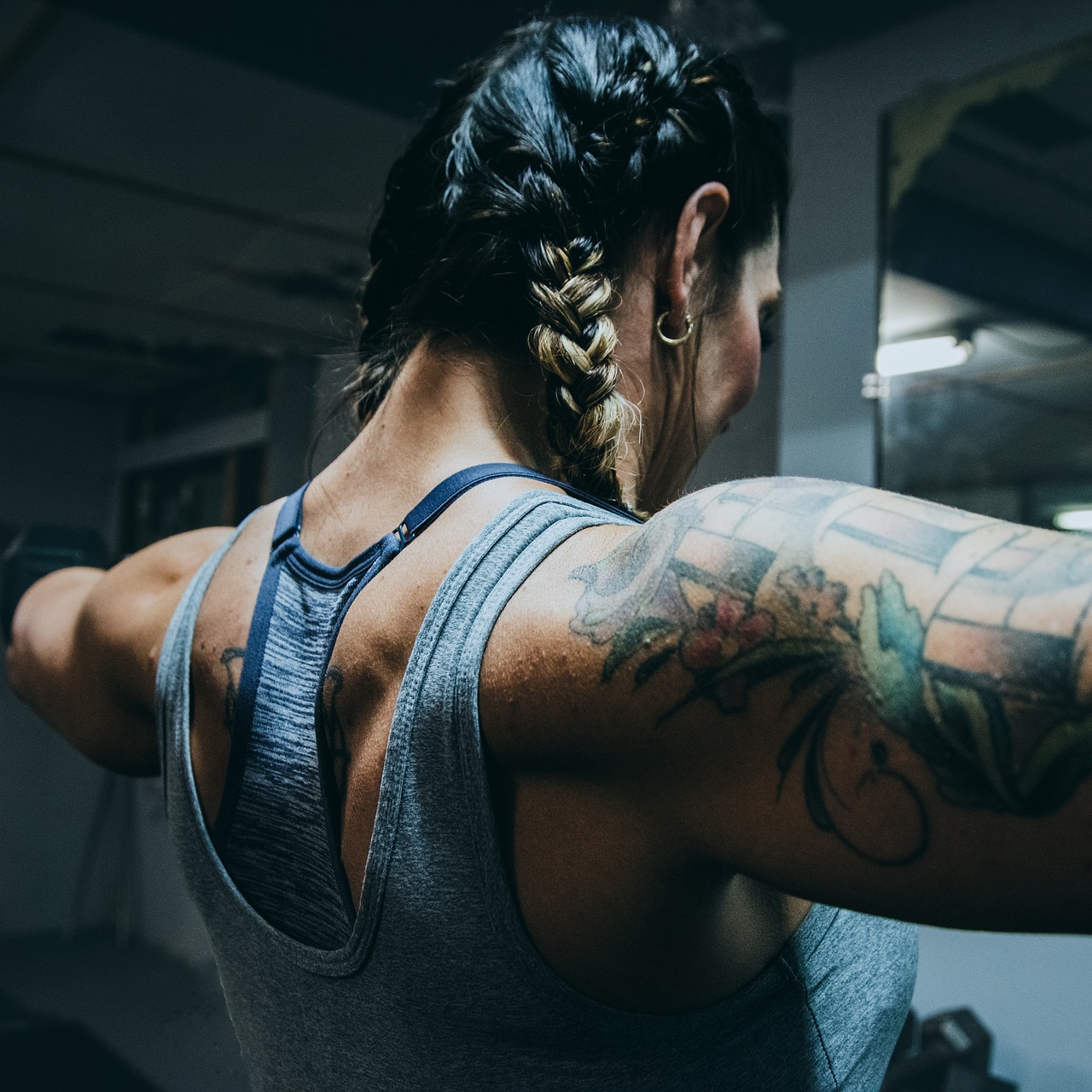 A young woman in a gym, with her back to the camera. She is wearing a grey tank top and has a tattoo on her left arm. Her hai...