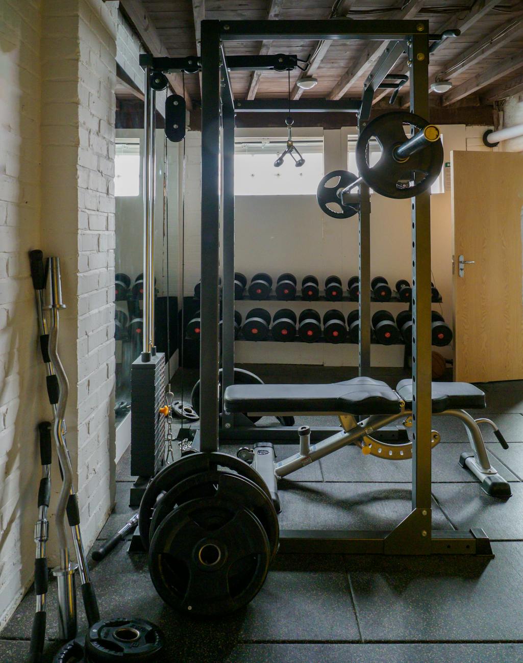 A gym with a weight rack and a bench. The weight rack is made of metal and has a barbell and weight plates attached to it. Th...