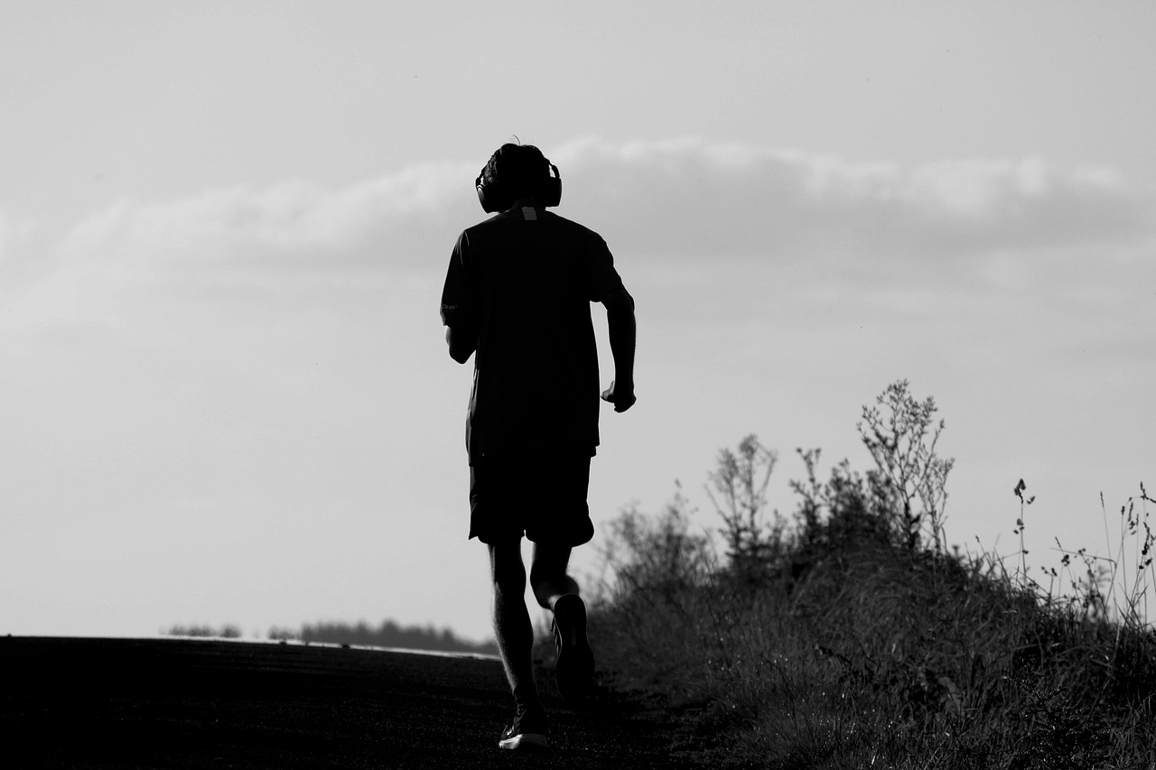 A black and white photograph of a person walking on a dirt path. The person is wearing a t-shirt, shorts, and sneakers. They ...