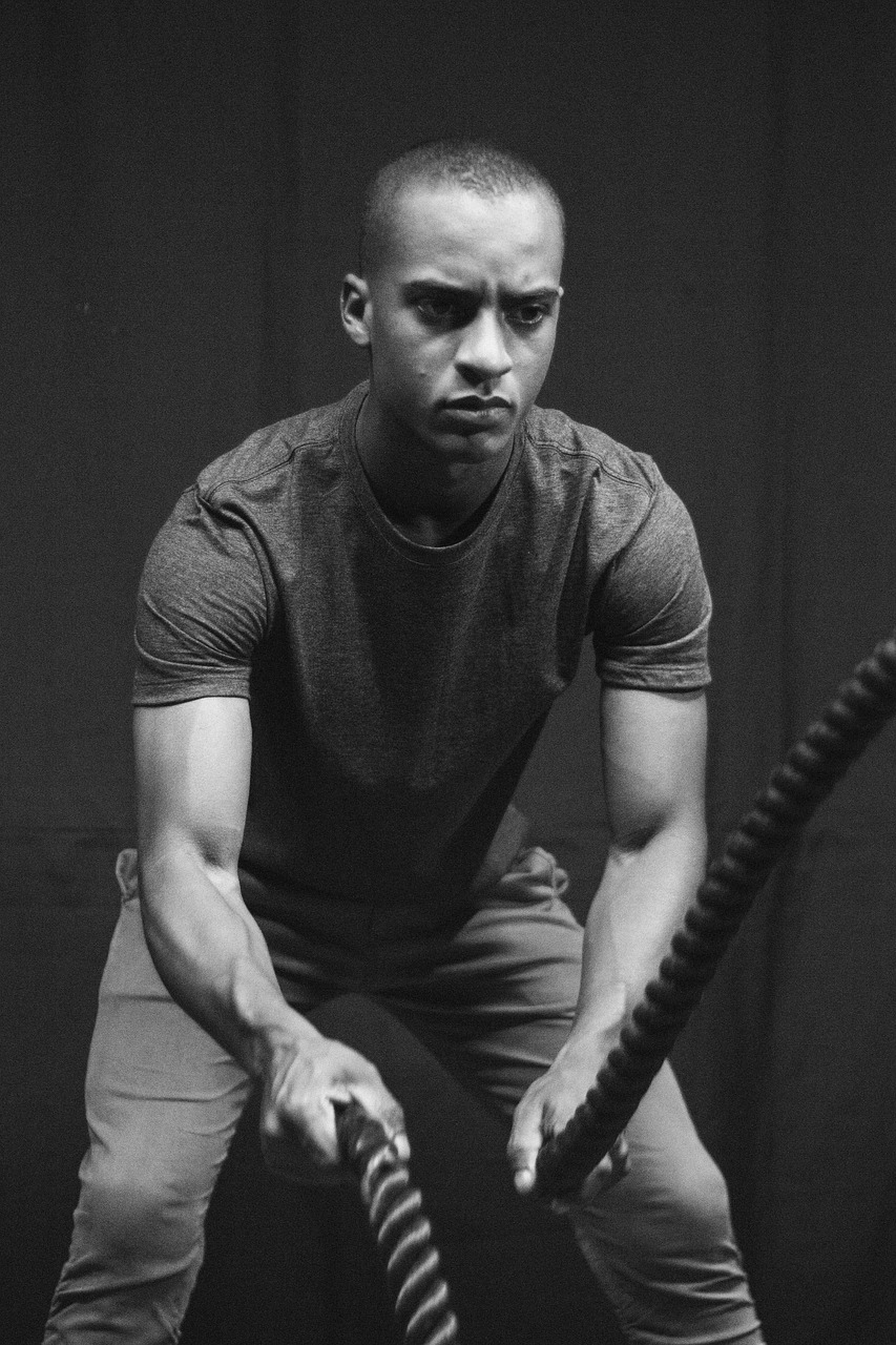 A black and white photograph of a young man in a gym. He is wearing a t-shirt and is holding a battle rope in his hands. The ...