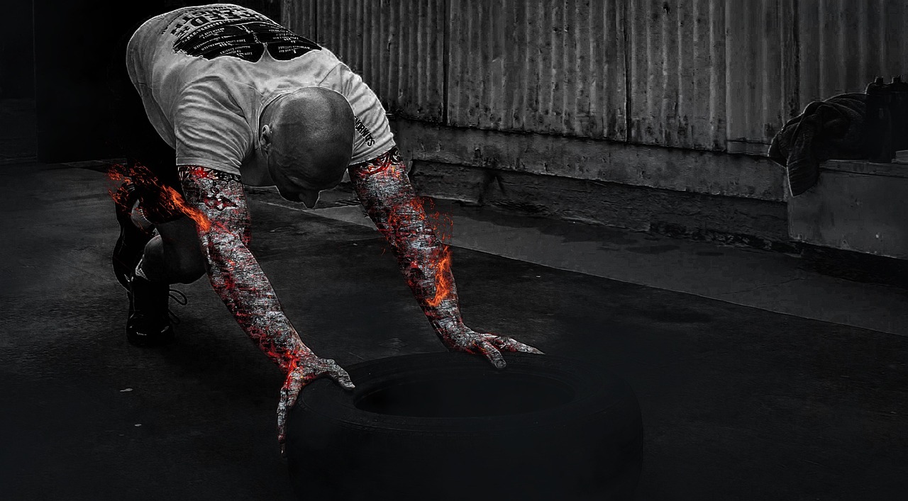 A man in a gym, performing push-ups on a black tire. He is wearing a white t-shirt with a graphic design on it and black shor...