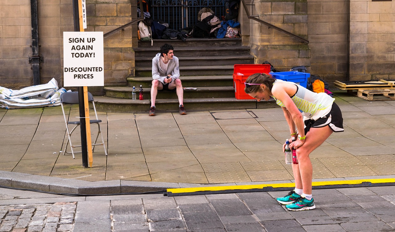 A young woman in a yellow tank top and black shorts bending over on a sidewalk. She is holding a water bottle in her right ha...