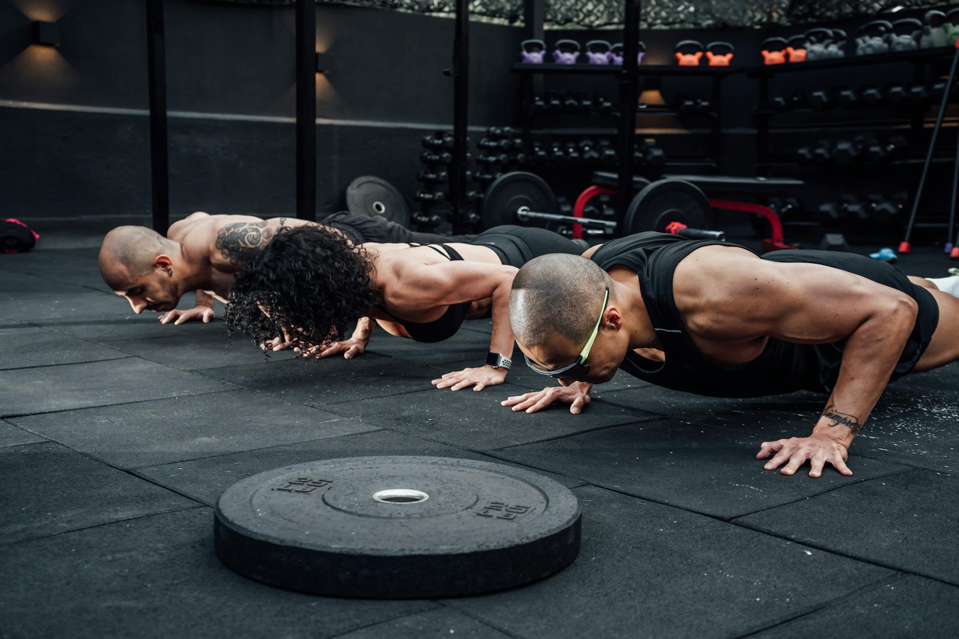 Three men doing push-ups in a gym. They are all wearing black workout clothes and are in a plank position with their hands an...