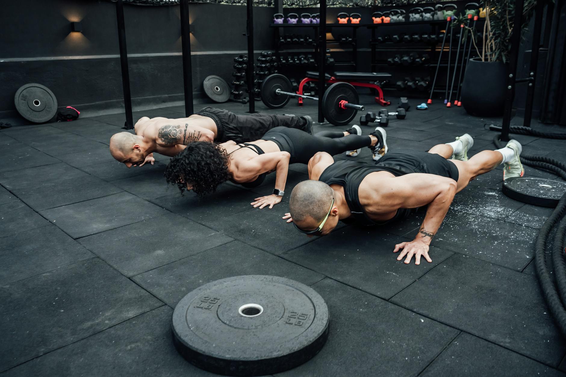 Three people performing push-ups in a gym. They are lying on their stomachs on the floor, with their arms and legs stretched ...