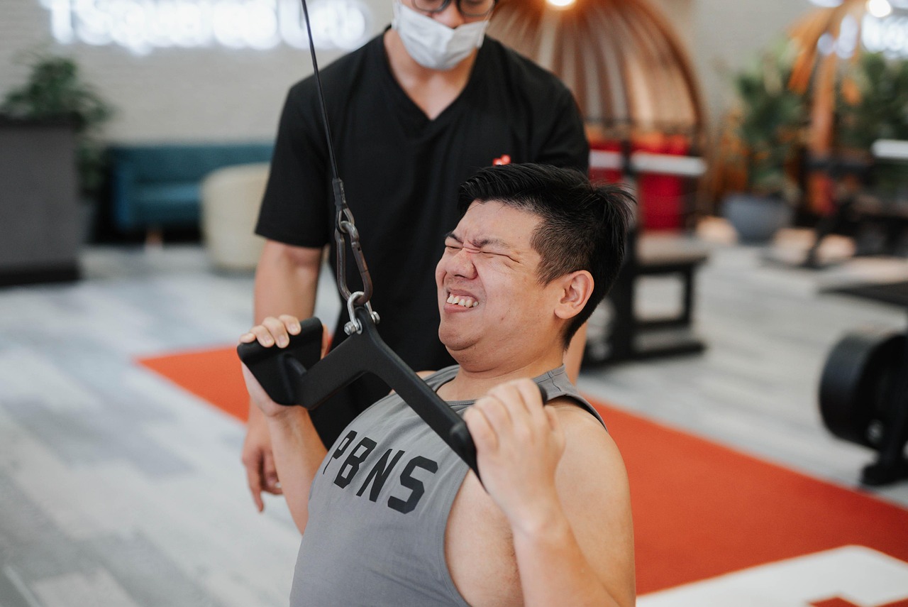 A young man in a gym, wearing a grey tank top with the word "PBS" written on it. He is holding onto a black resistance band w...