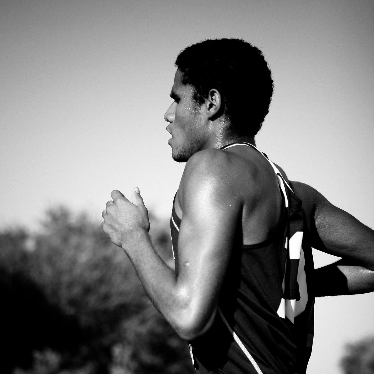 A black and white photograph of a young man running. He is wearing a sleeveless tank top and appears to be in motion, with hi...