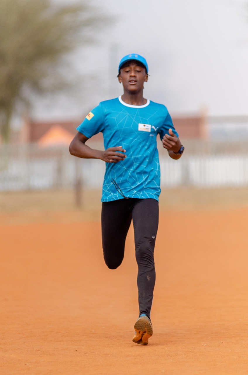 A young man running on an orange-colored track. He is wearing a blue t-shirt, black leggings, and a blue cap. He appears to b...