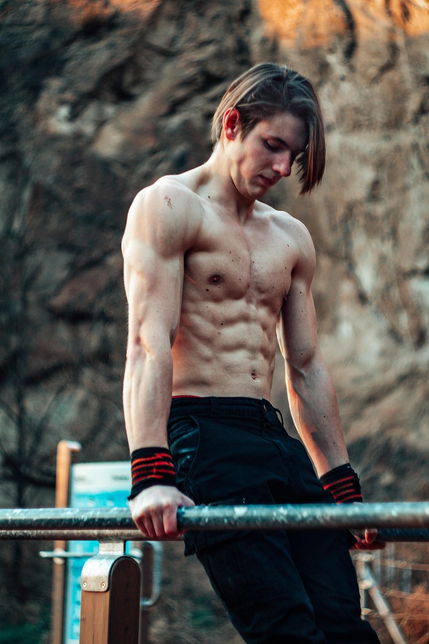 A young man standing on a horizontal bar in a gym. He is shirtless and wearing black shorts and red and black wristbands. His hair is styled in a messy yet fashionable manner and he is looking down at the bar with a serious expression on his face. The background is blurred, but it appears to be an outdoor setting with a rocky cliff and trees. The lighting is soft and warm, giving the image a dreamy and ethereal feel.