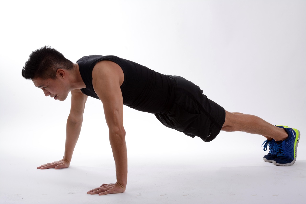 A young man performing a push-up exercise. He is wearing a black tank top and black shorts, and blue and yellow running shoes...