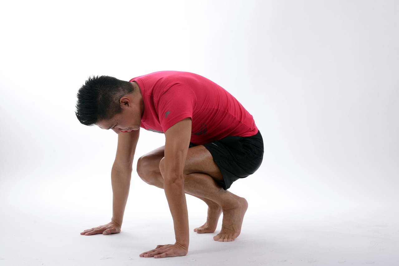 A young man in a pink t-shirt and black shorts performing a yoga pose. He is in a crouched position with his hands on the gro...