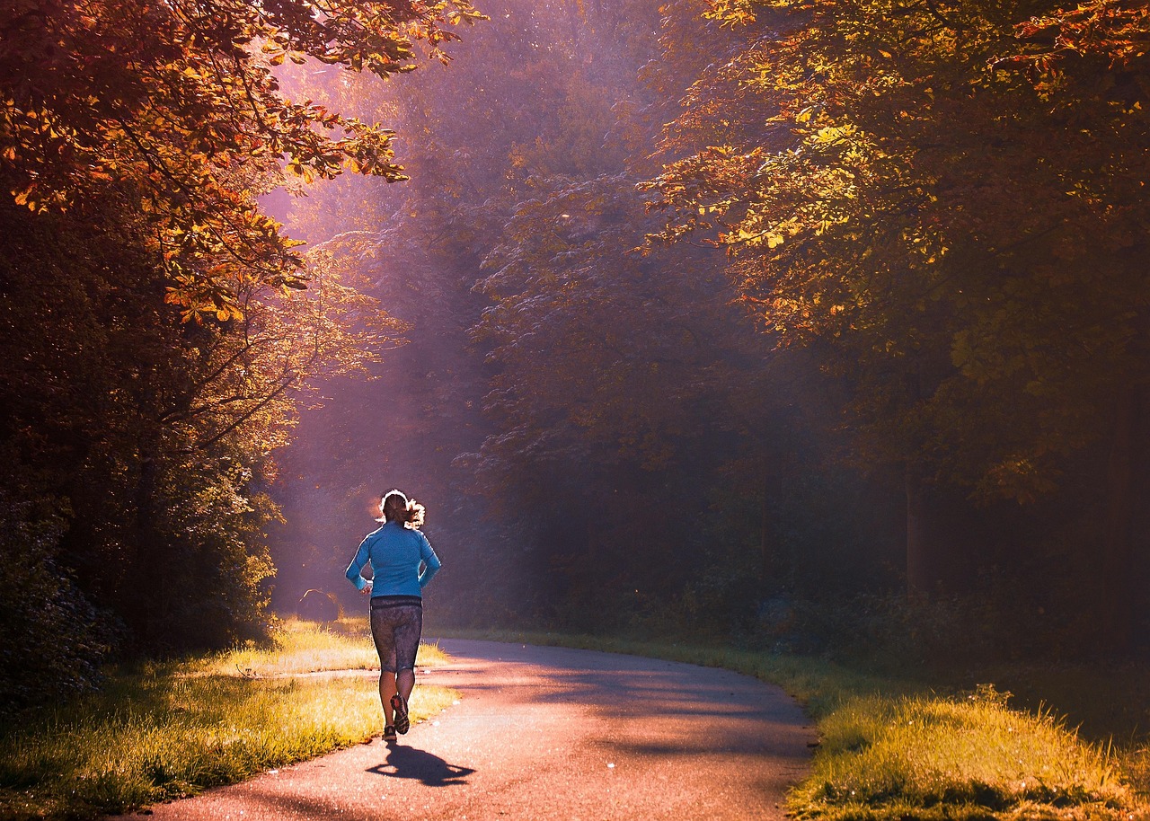 A person running on a paved path in a forest. The person is wearing a blue jacket and black shorts and is running towards the...