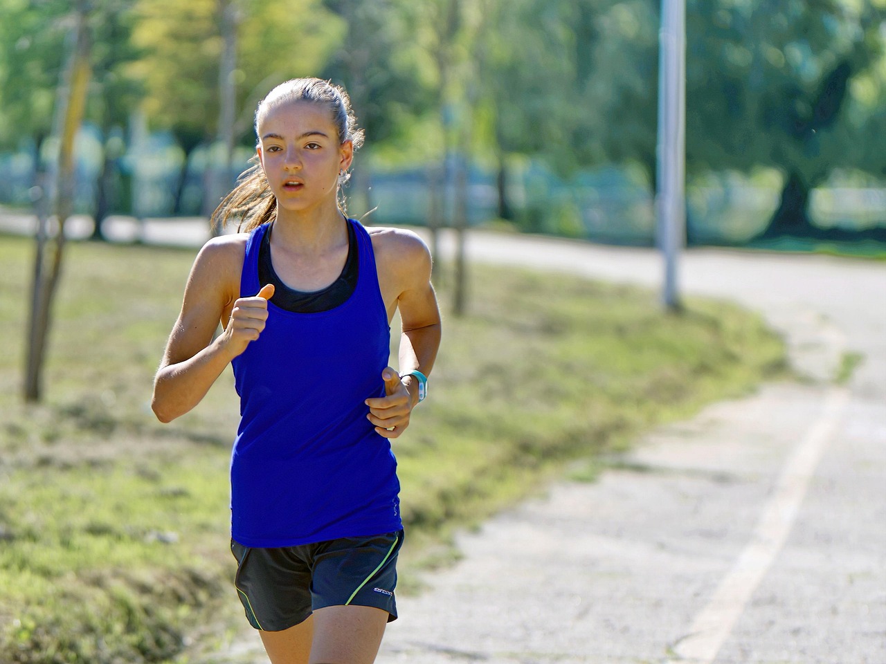A young woman running on a paved path in a park. She is wearing a blue tank top and black shorts. Her hair is pulled back in ...
