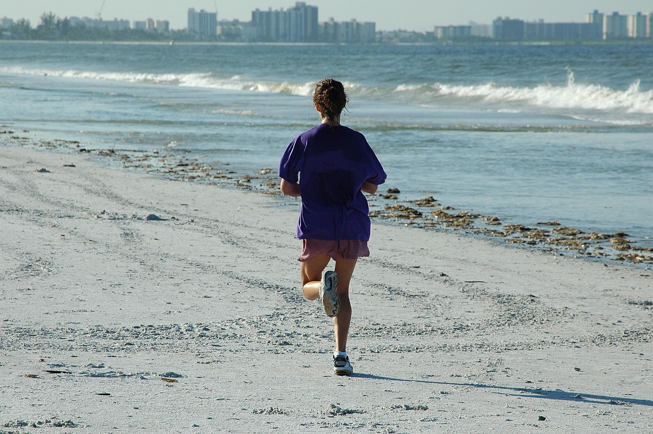 A person running on a sandy beach. The person is wearing a purple t-shirt and shorts and is running towards the ocean. The ocean is visible on the right side of the image, with small waves crashing onto the shore. In the background, there are several buildings and skyscrapers visible. The sky is blue and the weather appears to be sunny and warm.