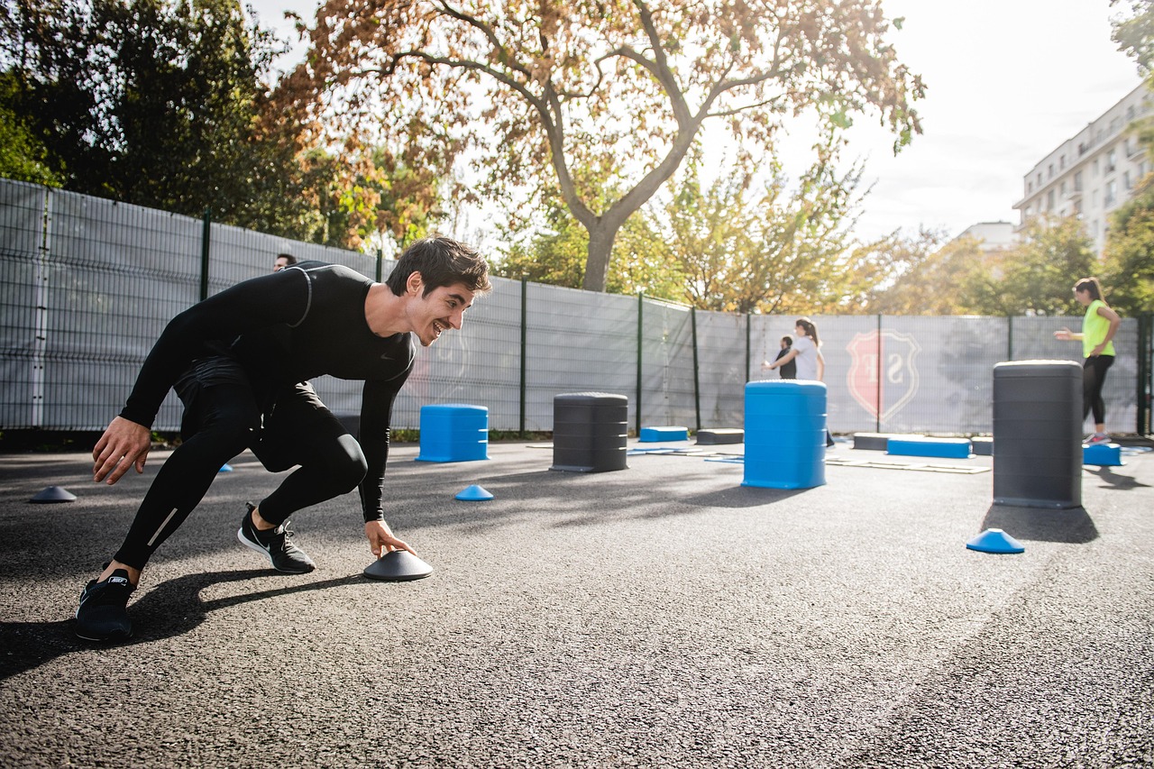 A young man in a black workout outfit, crouching down on a concrete surface with blue cones scattered around him. He is in a ...