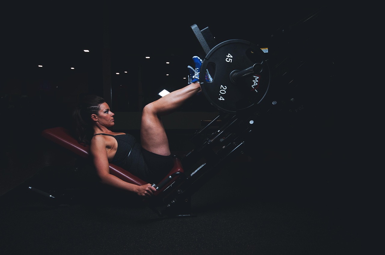 A young woman performing a leg press exercise on a bench press machine in a gym. She is wearing a black tank top and shorts and is lying on her back with her legs bent at the knees and her feet flat on the bench. The machine is black and has a barbell attached to it. The woman is holding onto the barbell with both hands and appears to be in the middle of a workout. The background is dark and the lighting is dim, suggesting that the gym is dimly lit. The image is taken from a low angle, focusing on the woman and the machine.