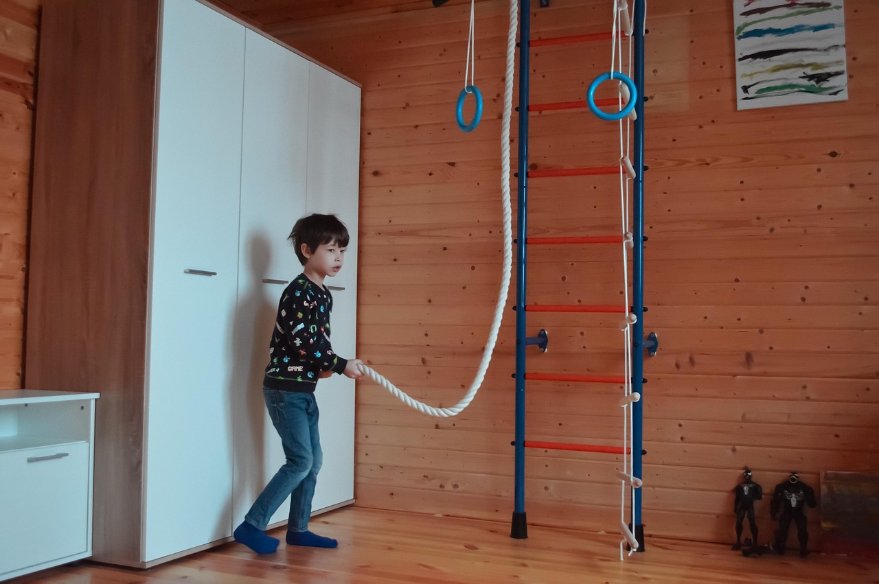 A young boy playing with a rope in a room with wooden walls. He is standing in front of a wooden ladder with blue rings hangi...