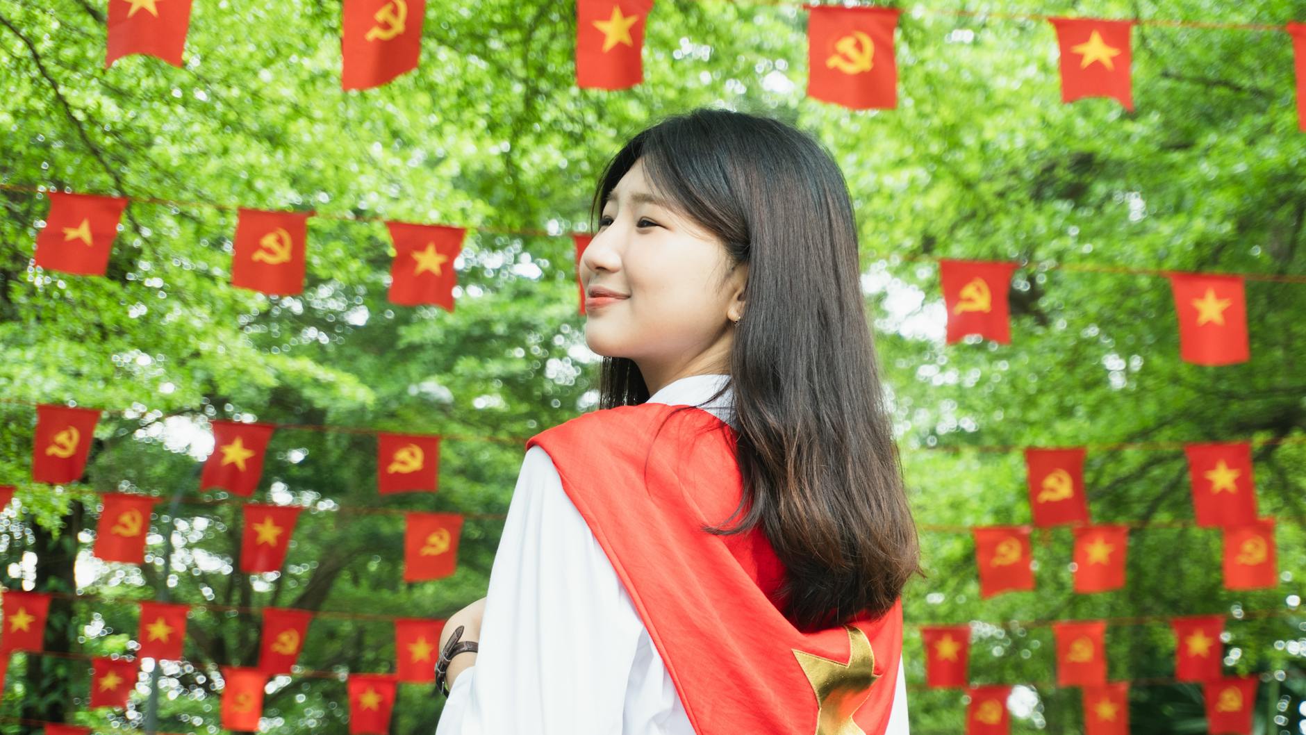 A young woman standing in front of a background of red Chinese flags hanging from a string. She is wearing a white blouse wit...