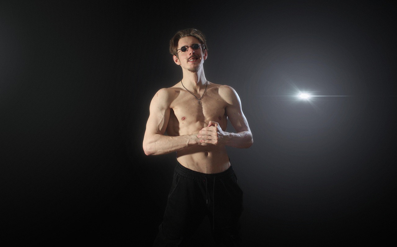 A young man standing in a dark room with a bright light shining on him. He is shirtless and wearing black sweatpants and sung...