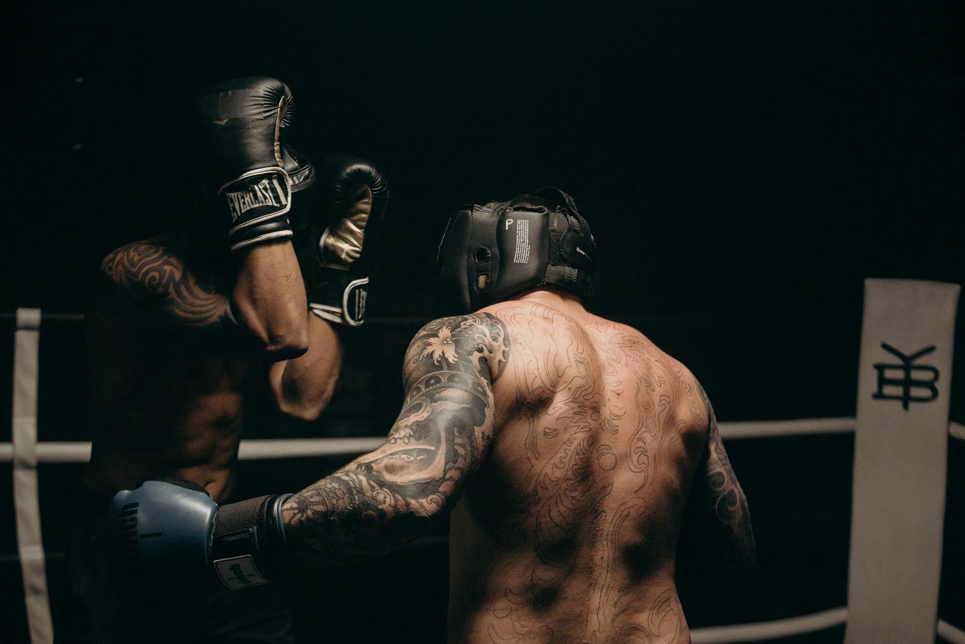 Two men in a boxing ring, both wearing black boxing gloves and headgear. The man on the left is throwing a punch at the other man, who is standing in front of him with his back to the camera. Both men have tattoos on their arms and back. The background is dark and the ring is surrounded by ropes. The image is taken from a low angle, looking up at the two men. The overall mood of the image is intense and focused.