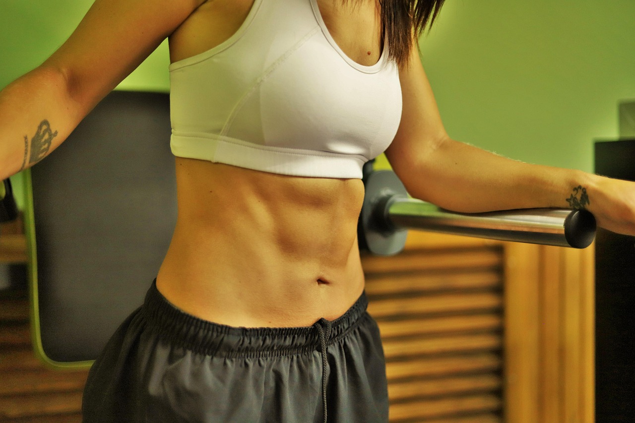 A young woman in a gym, wearing a white sports bra and black shorts. She is standing in front of a green wall and is holding ...