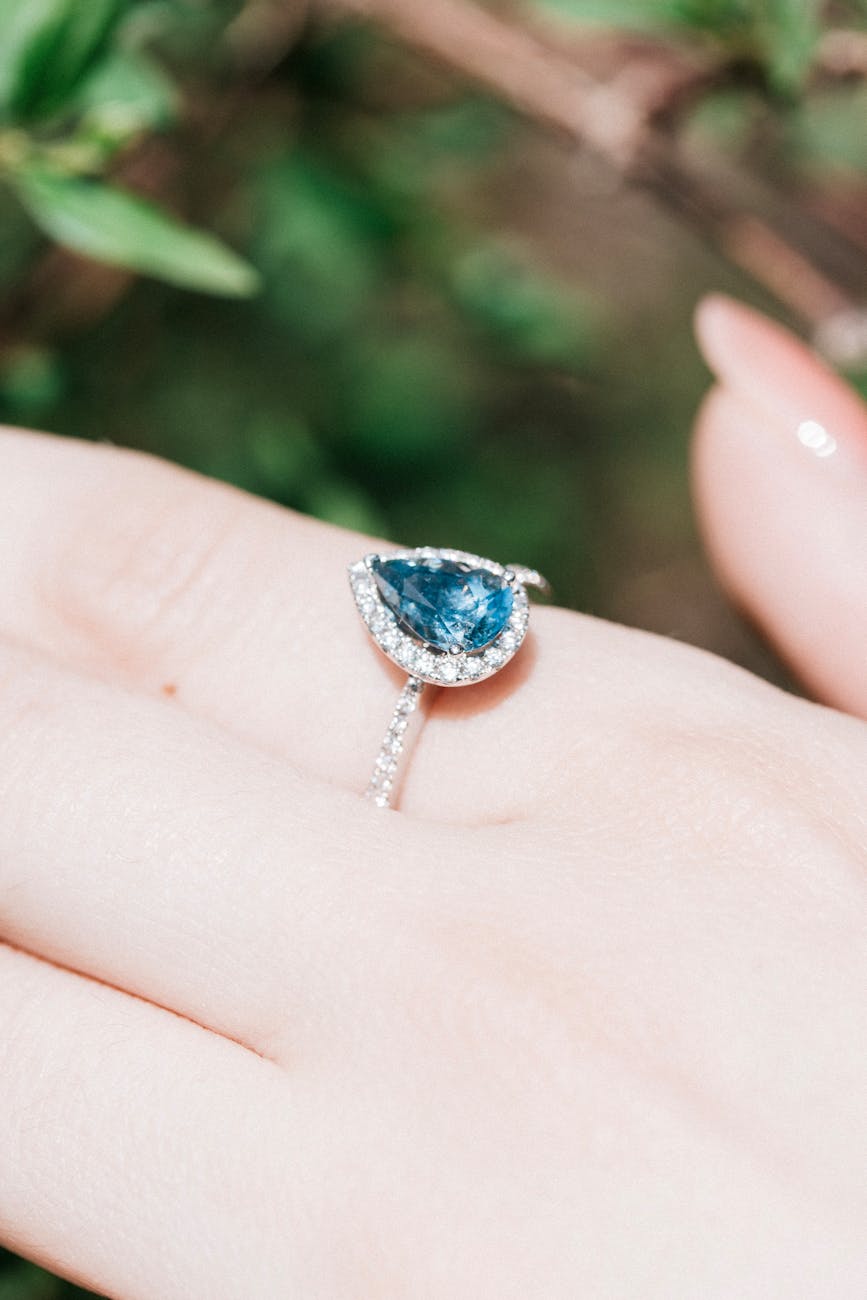A close-up of a person's hand with a ring on their ring finger. The ring has a large, oval-shaped blue gemstone in the center...