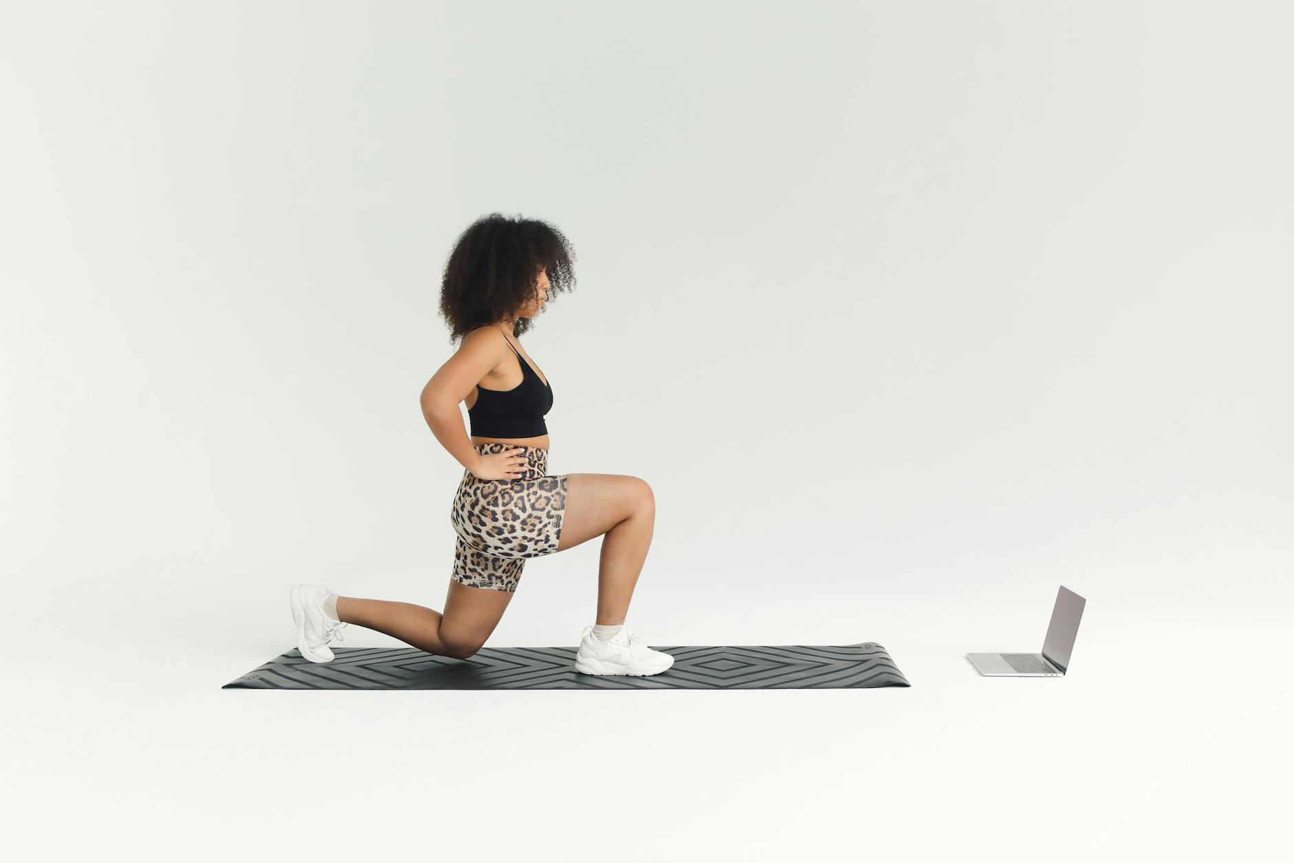 A young woman in a black sports bra and leopard print shorts performing a lunge exercise on a black yoga mat. She is in a sid...