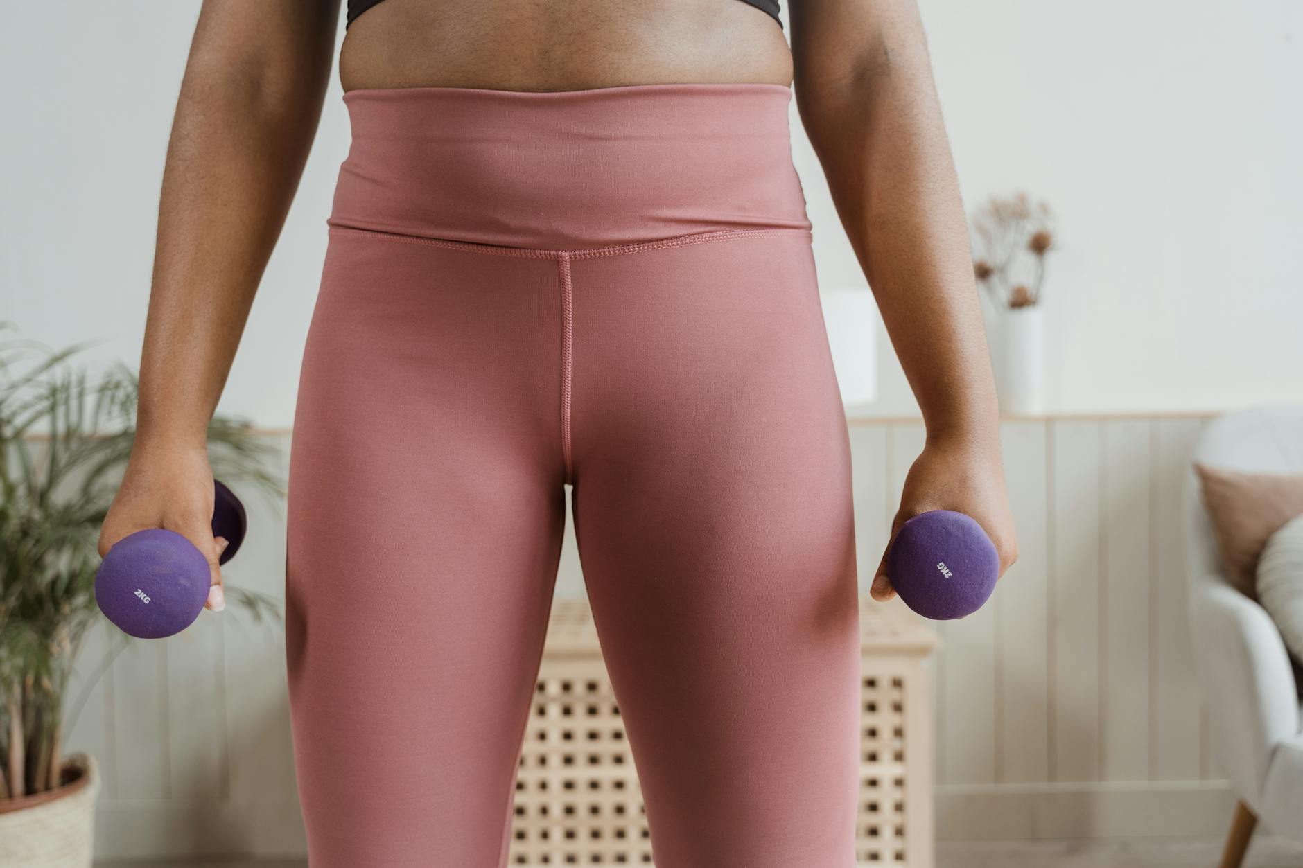 The lower body of a woman from the waist down, holding two purple dumbbells in her hands. She is wearing a black sports bra a...
