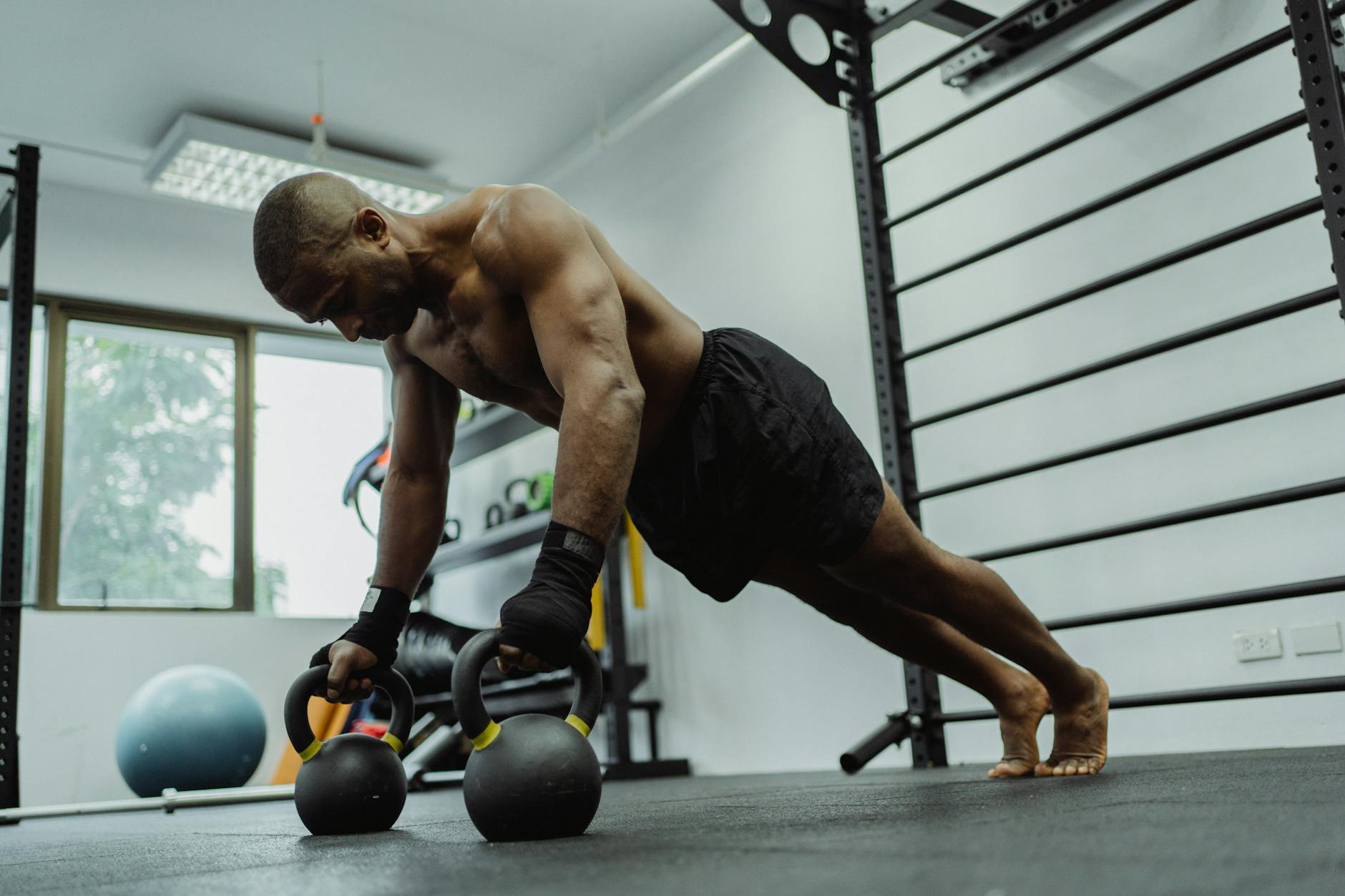 A man performing a push-up exercise with kettlebells in a gym. He is wearing black shorts and black gloves, and his body is in a plank position with his feet shoulder-width apart and his arms extended above his head. His hands are holding the kettlebell handles with both hands and his feet are planted firmly on the ground. The background shows a gym equipment rack and a window with a view of trees outside. The man appears to be focused and determined as he performs the exercise.