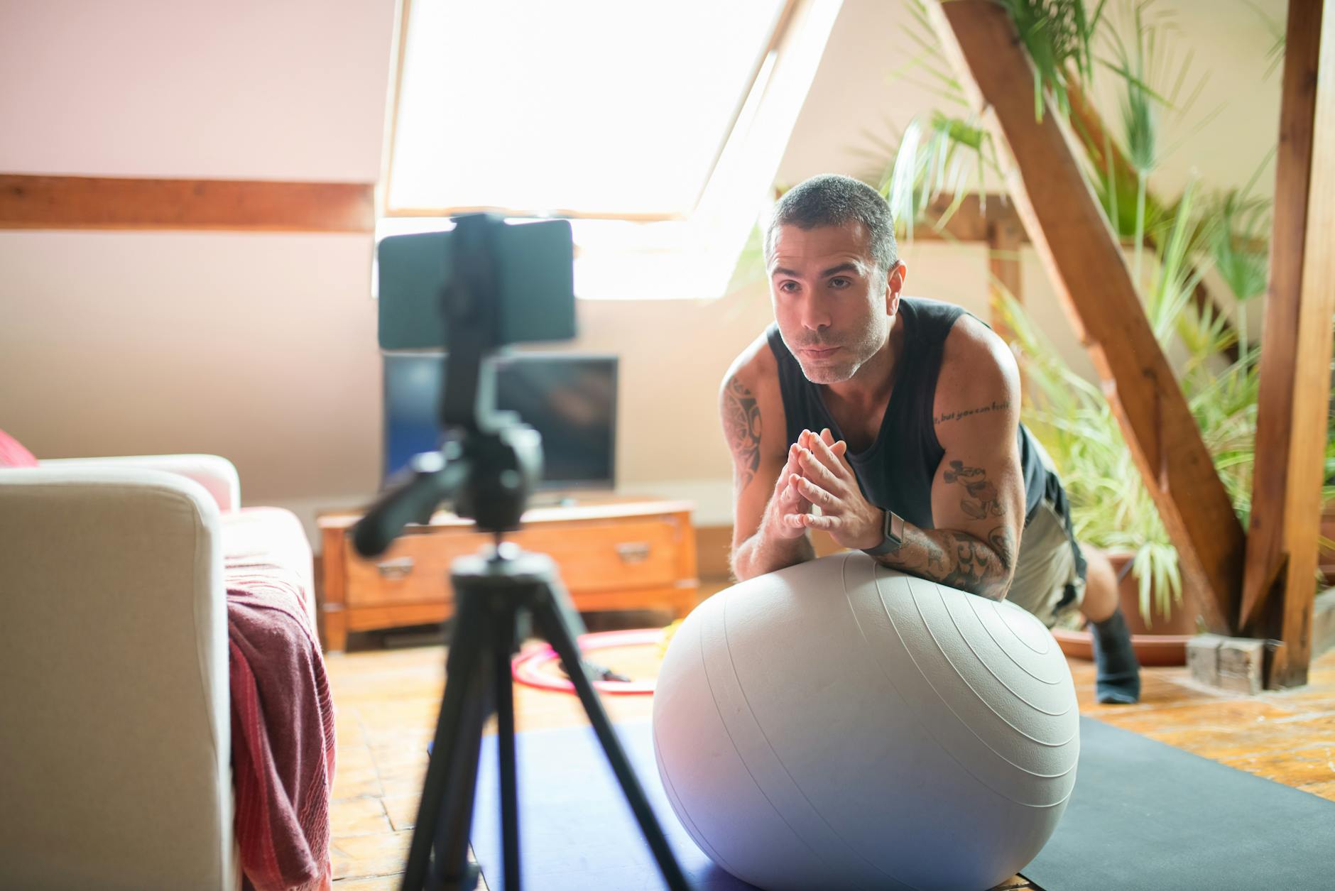 A man sitting on an exercise ball in a living room. He is wearing a black tank top and has tattoos on his arms and chest. He ...