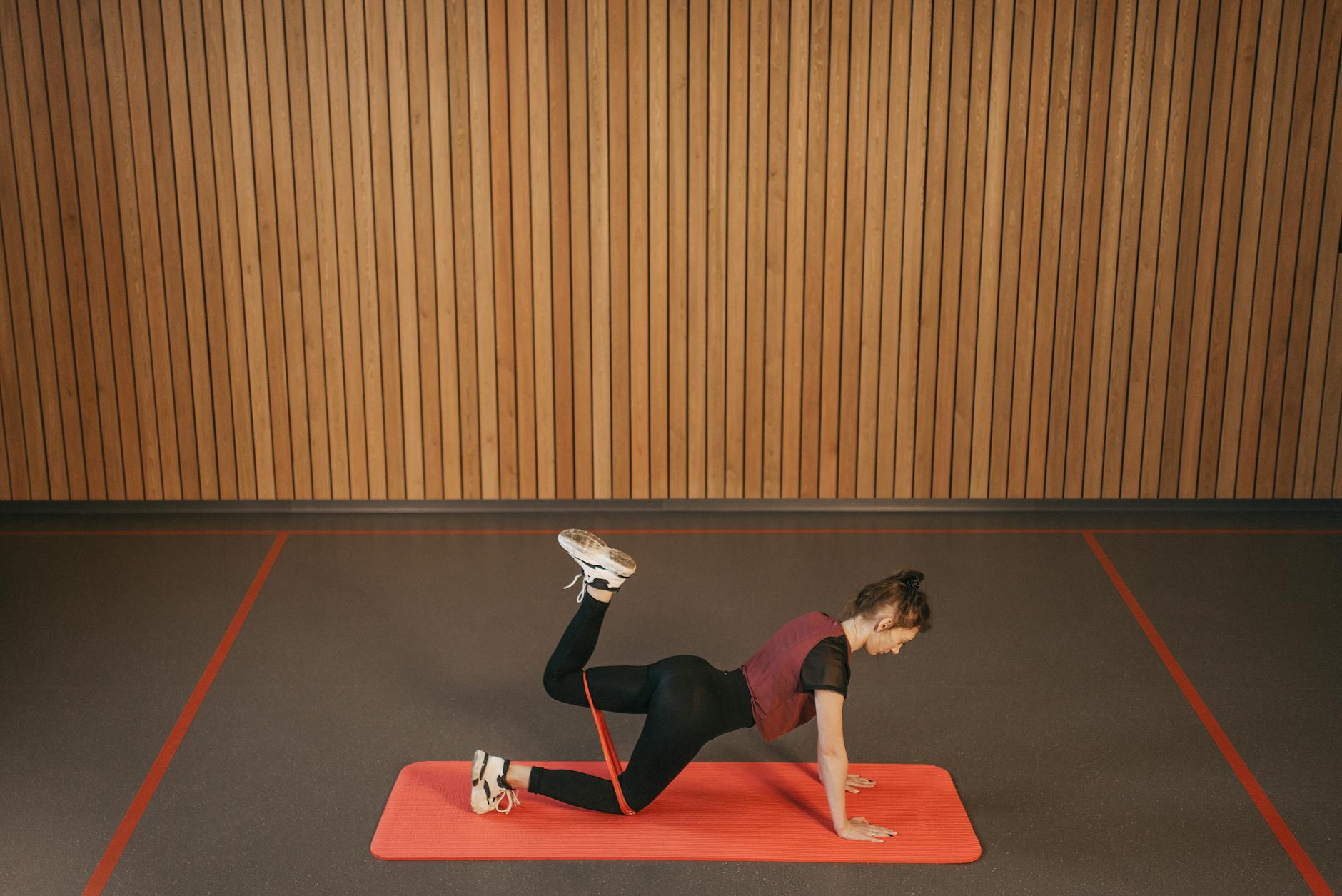A young woman performing a plank exercise on a red mat in a gym. She is wearing a maroon tank top, black leggings, and white ...