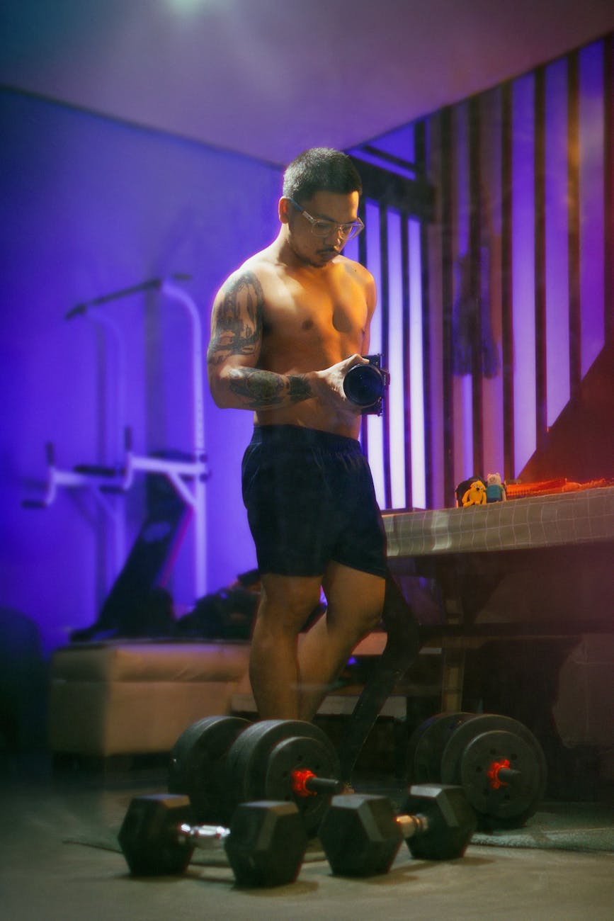 A young man in a gym, standing in front of a table with dumbbells on it. He is shirtless and wearing black shorts, and has tattoos on his arms and chest. He appears to be in the middle of a workout, as he is holding a dumbbell in his hands and is looking down at it with a focused expression on his face. The room is dimly lit with purple and blue lights, and there are various exercise equipment in the background. The overall mood of the image is intense and focused.