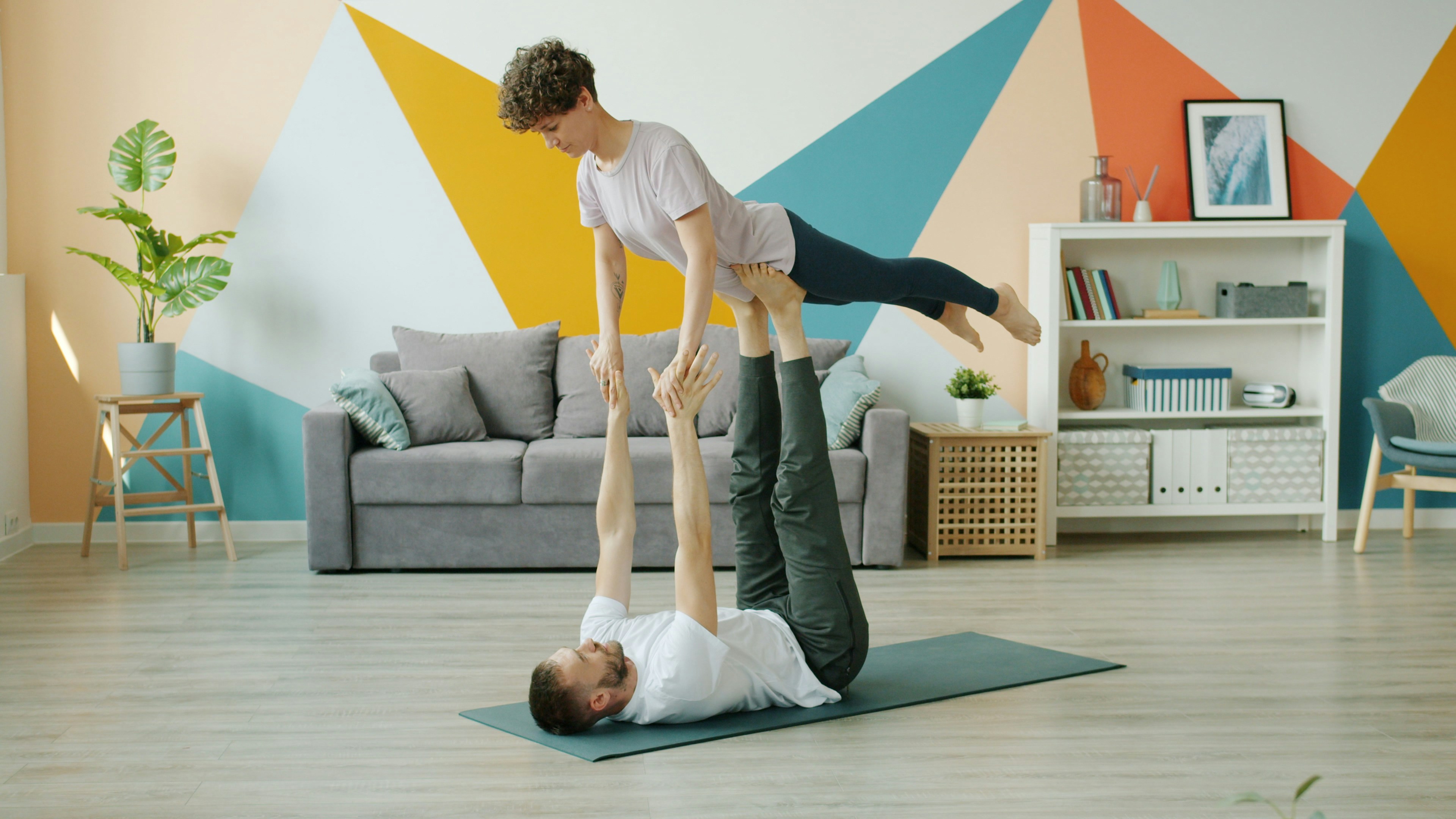 A man and a woman performing a yoga pose in a living room. The man is lying on his back on a green yoga mat, with his legs st...