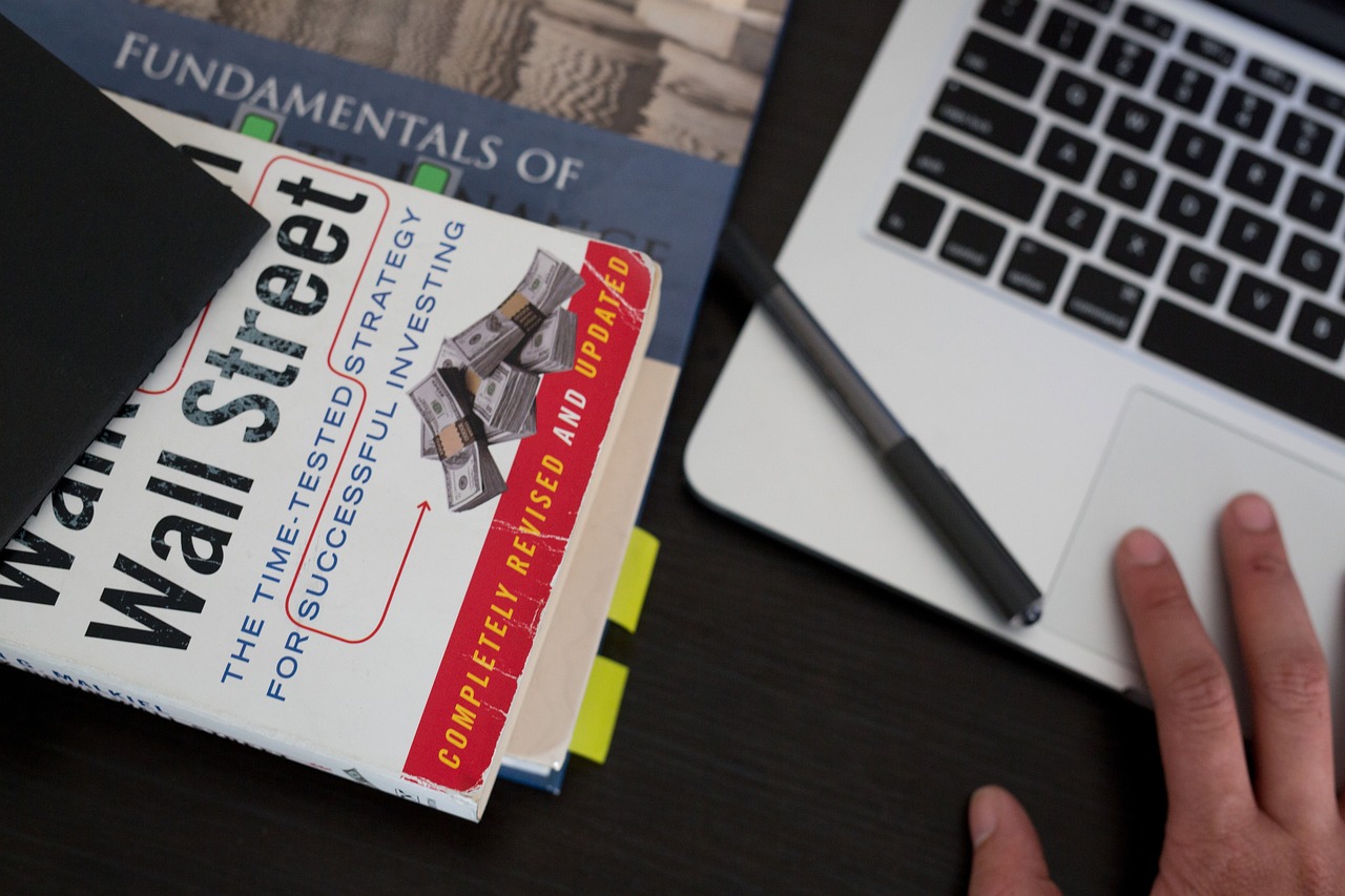 A desk with a laptop, a notebook, and a pen on it. On the desk, there is a book titled "Fundamentals of Wall Street: The Time-Tested Strategy for Successful Investing" with a red cover and white text. The book appears to be a comprehensive guide to financial planning and investing. A person's hand is visible on the right side of the image, resting on the desk.