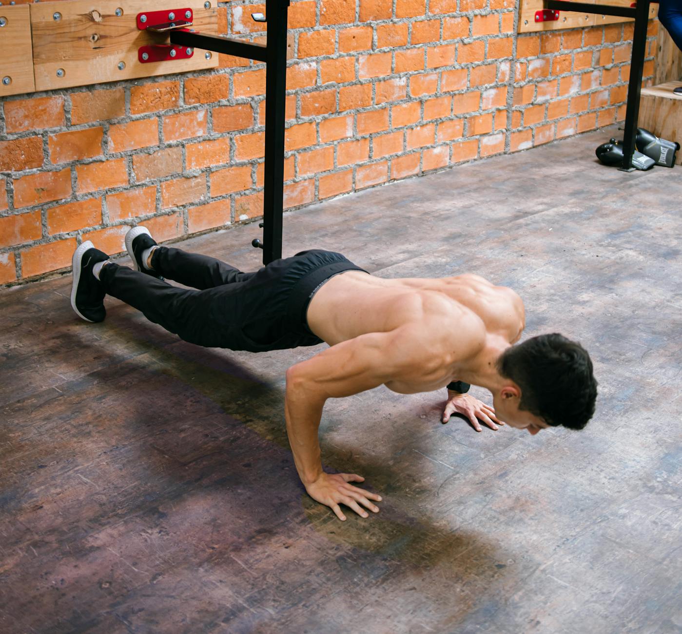A young man performing a push-up exercise in a gym. He is wearing black shorts and white sneakers. His body is in a plank pos...