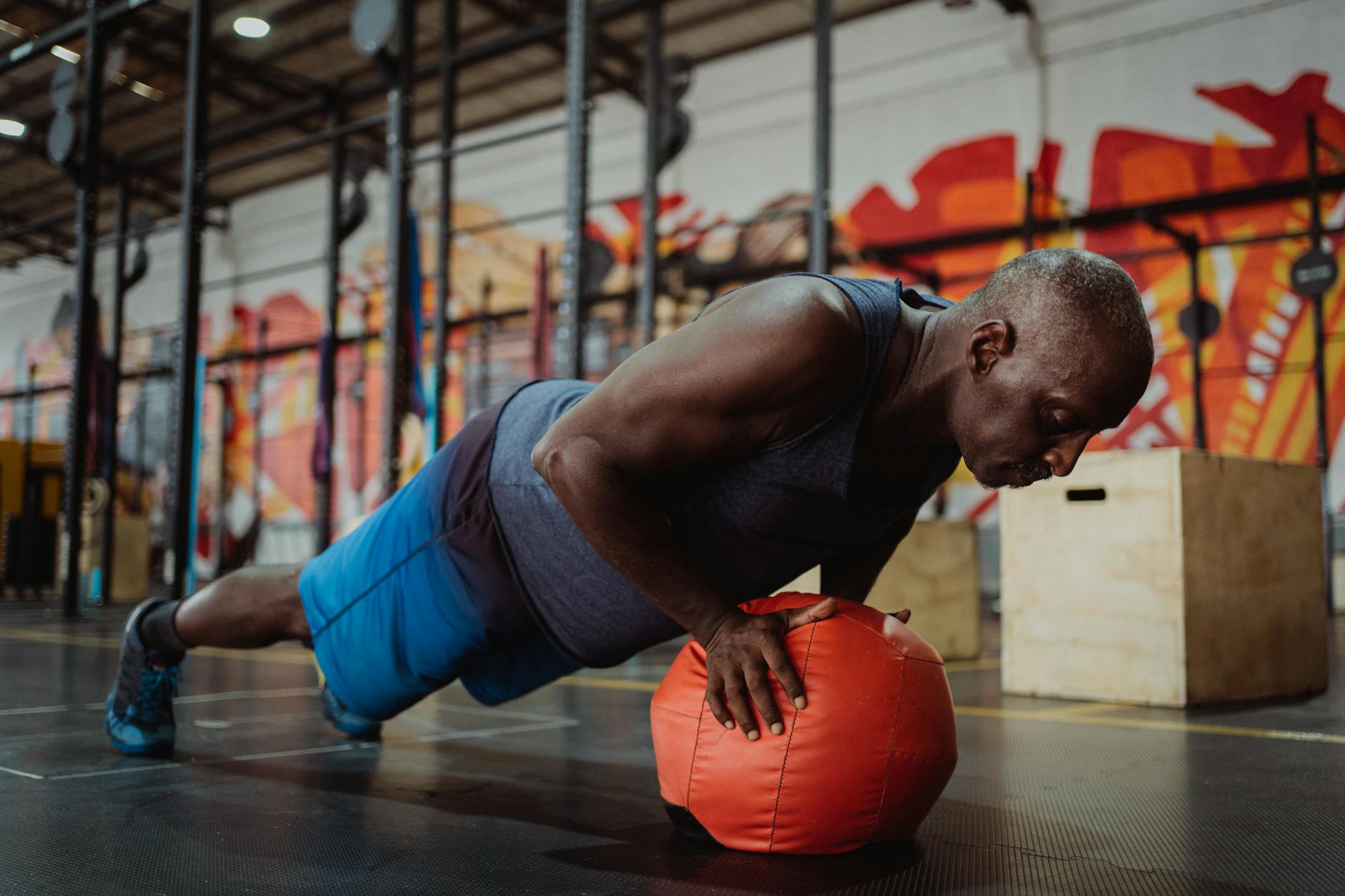 A man in a gym, performing a push-up exercise with a red medicine ball. He is wearing a blue tank top and blue shorts, and hi...