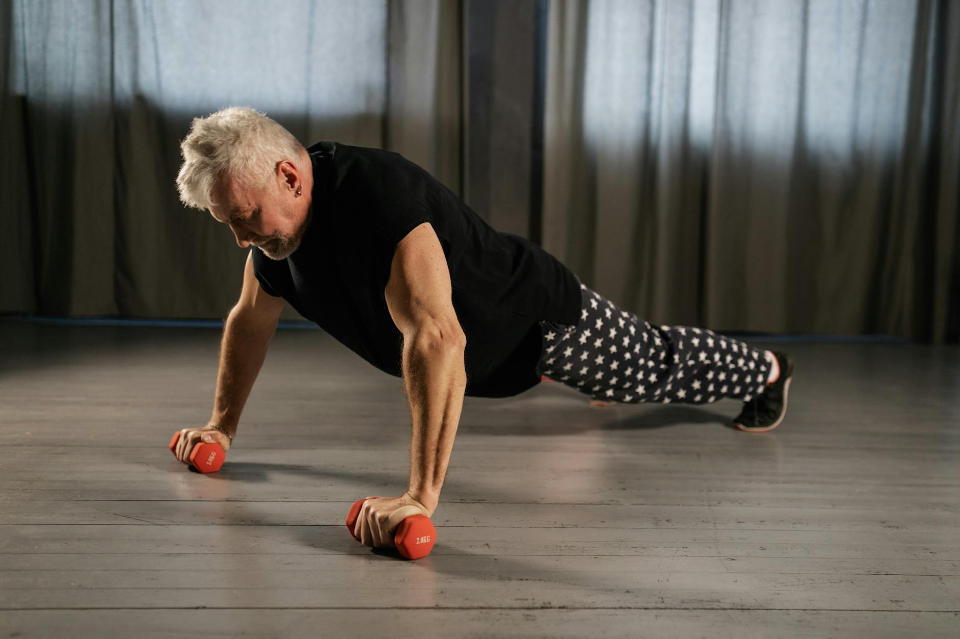 An elderly man performing a push-up exercise with red dumbbells. He is wearing a black t-shirt and patterned pants. The man i...