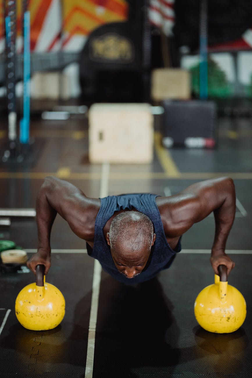 A man performing a push-up exercise with two yellow kettlebells. He is in a gym with a black mat on the floor and various gym...