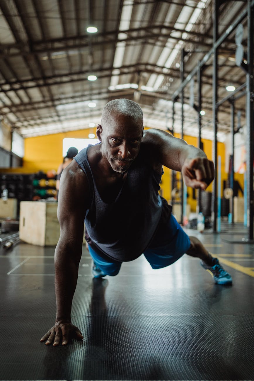 A man in a gym, performing a push-up exercise. He is wearing a black tank top and blue shorts, and his body is in a plank pos...