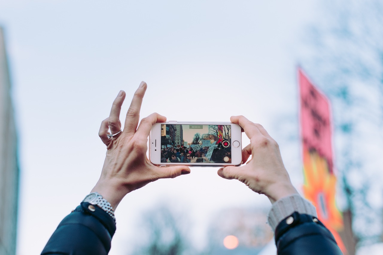 A person's hands holding a white iPhone with their fingers up in the air, taking a photo of a crowd of people on a street. Th...