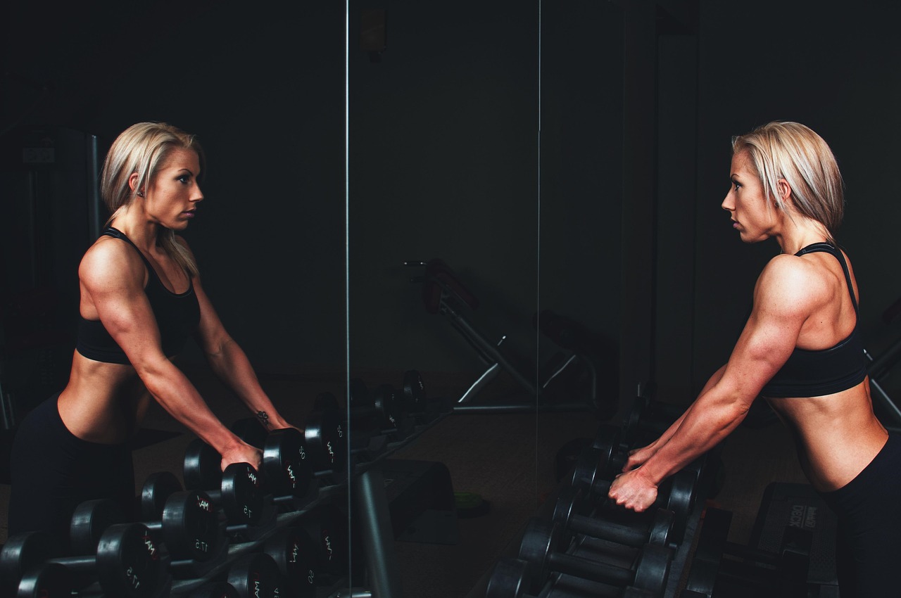 A young woman working out with dumbbells in a gym. She is wearing a black sports bra and black leggings and is standing in fr...