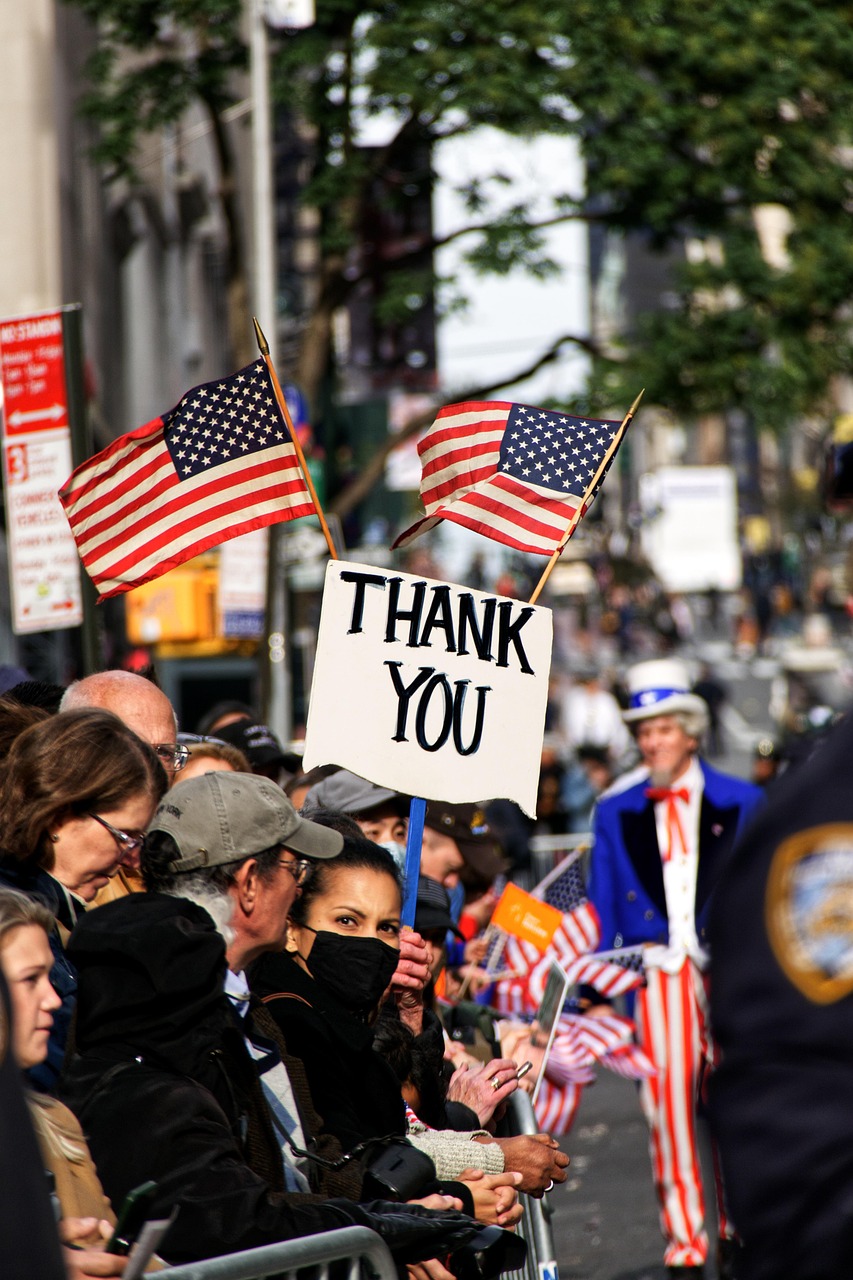 A crowd of people gathered on a street, holding American flags and a sign that reads "Thank You." The people appear to be par...