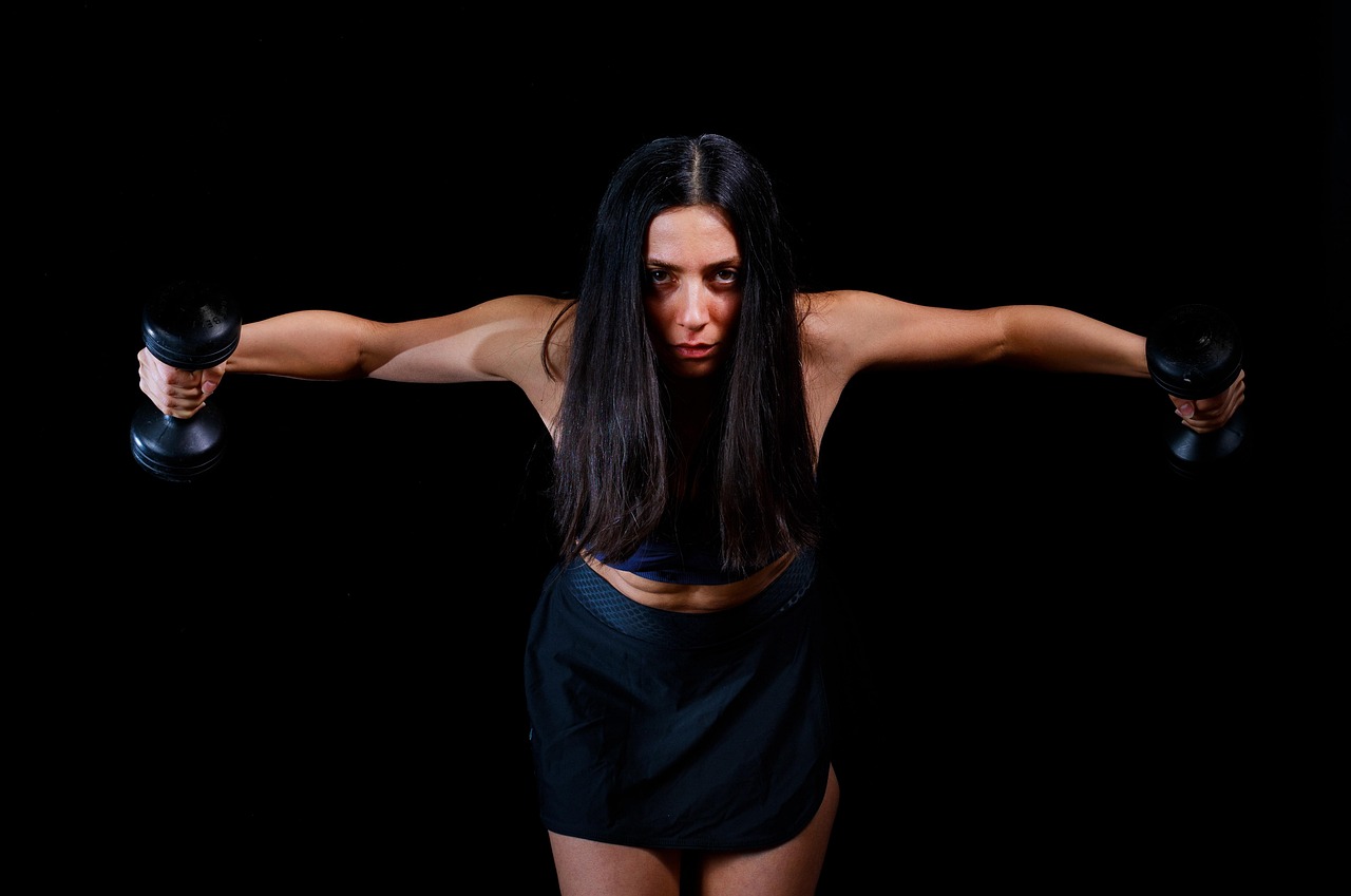 A young woman with long dark hair, wearing a black sports bra and shorts. She is standing in front of a black background and is holding two black dumbbells in her hands. The woman appears to be in the middle of a workout, with her arms stretched out to the sides and her head tilted downwards. She has a serious expression on her face and is looking directly at the camera. The lighting is dramatic and the overall mood of the image is intense.