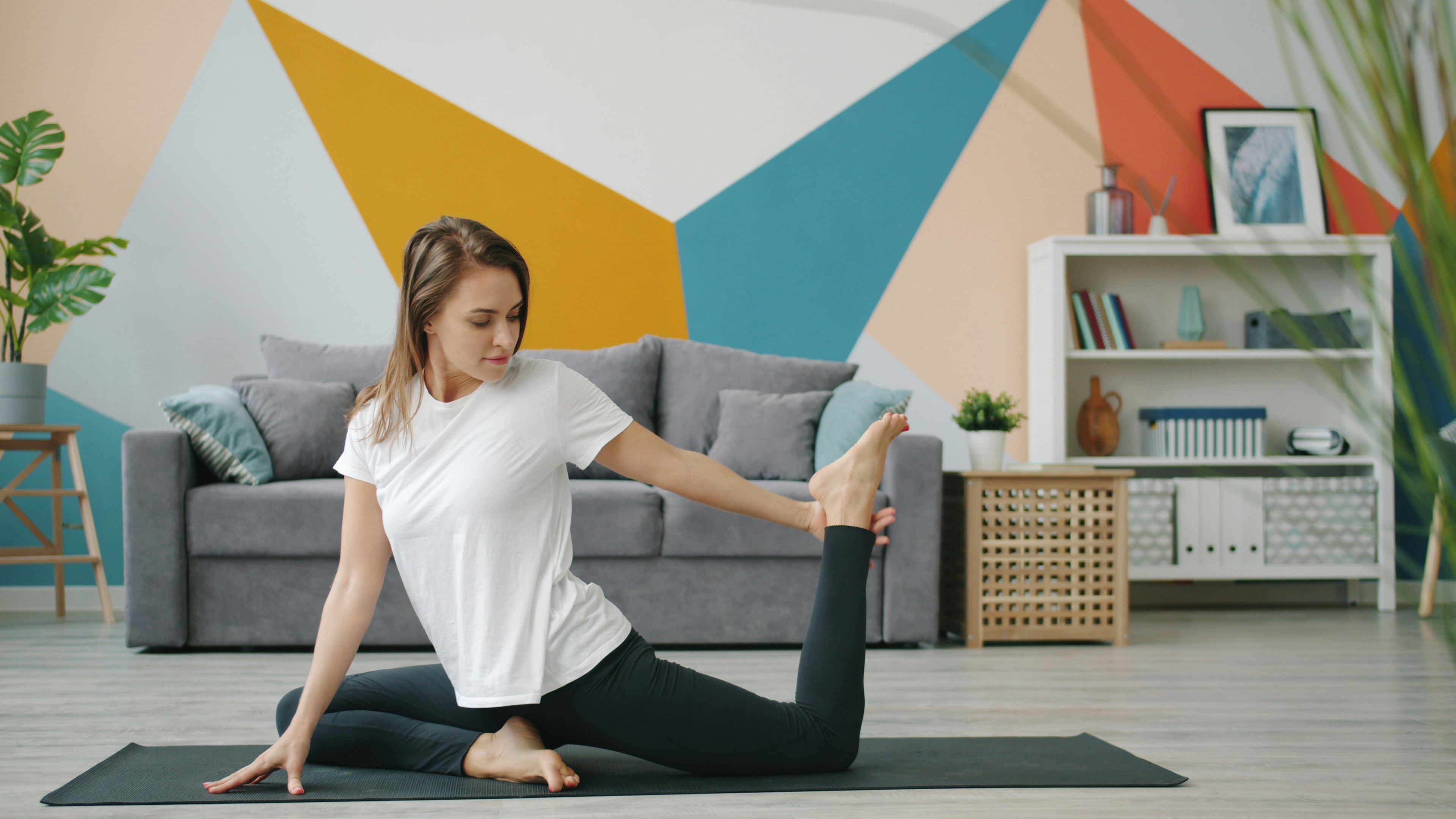 A young woman in a yoga pose on a black mat in a living room. She is wearing a white t-shirt and black leggings and is in a s...