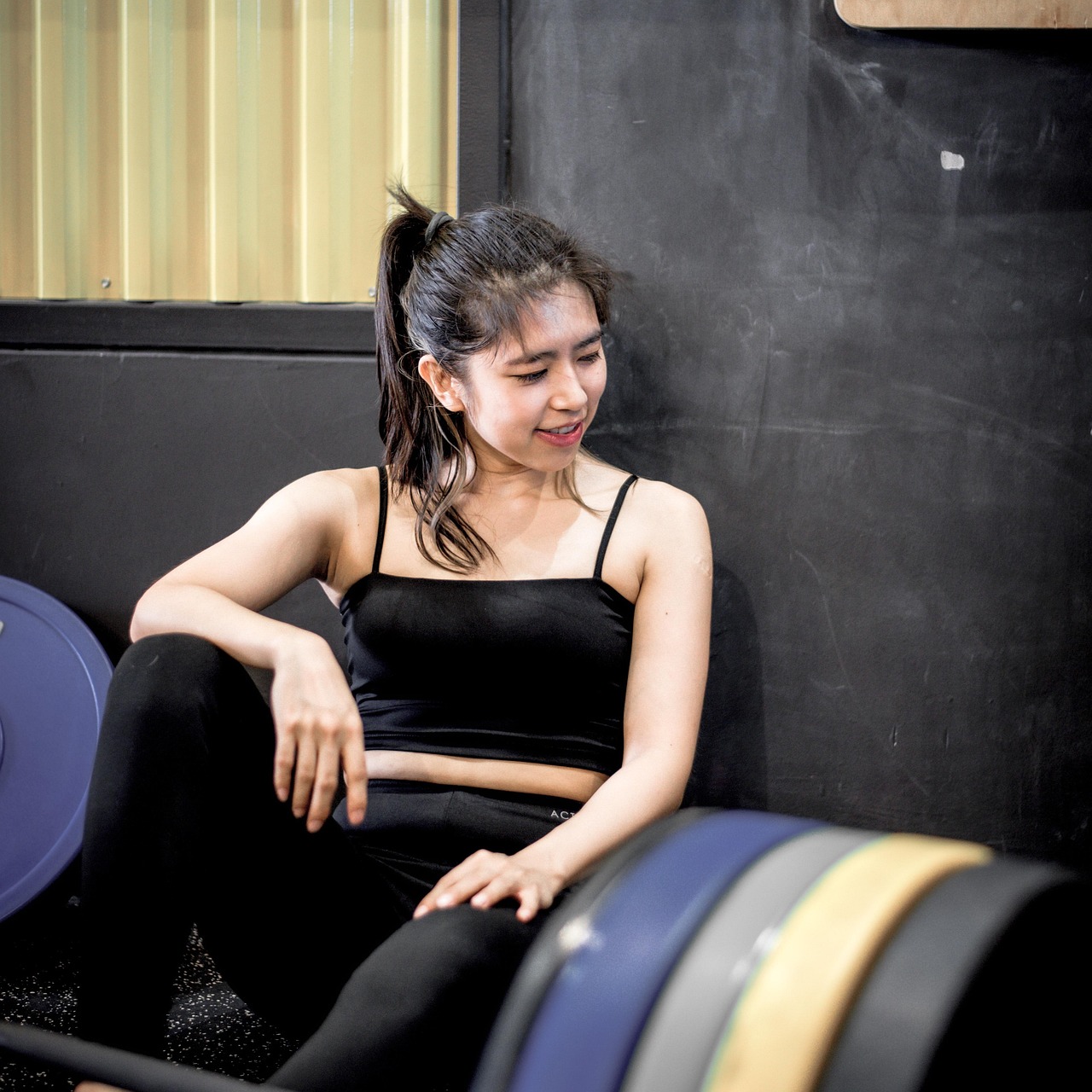 A young woman sitting on a bench in a gym. She is wearing a black crop top and black leggings. Her hair is pulled back in a p...