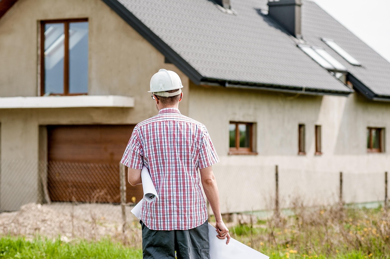 A man wearing a white hard hat and a red plaid shirt, walking towards a house under construction. He is holding a blueprint in his hand and appears to be walking towards the house. The house is a two-story building with a sloping roof and a garage on the left side. The exterior walls are painted in a light beige color and there is a fence on the right side of the image. The sky is overcast and there are some plants and grass in the foreground.