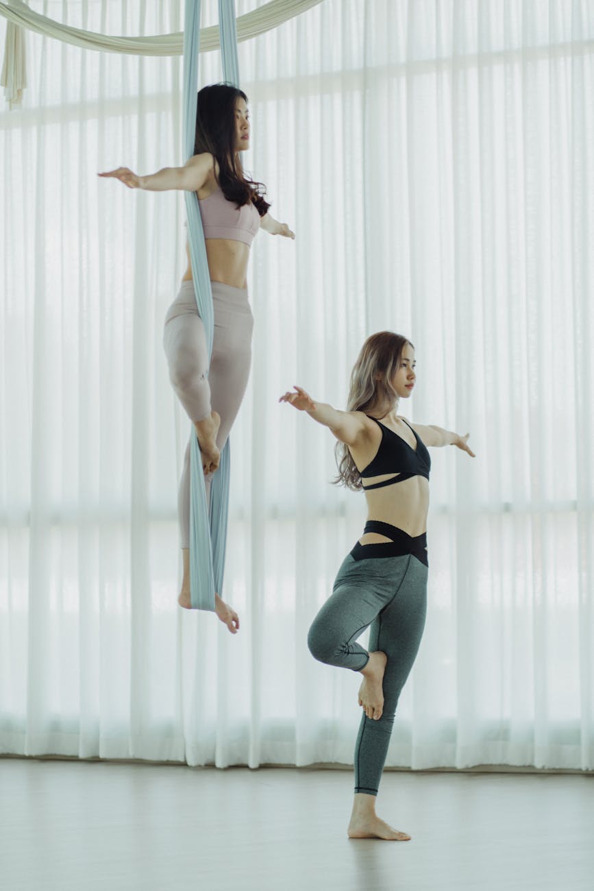 Two young women performing aerial acrobatics in a large room with white curtains. The woman on the left is wearing a pink spo...