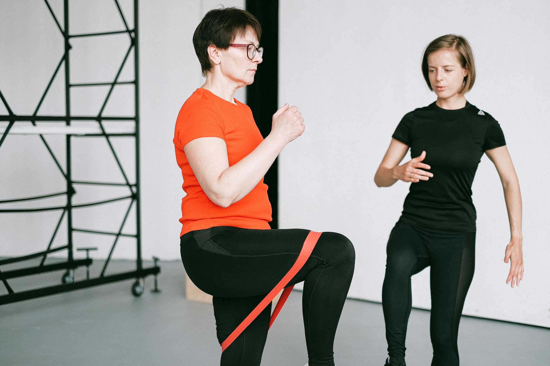 Two women in a dance studio. The woman on the left is wearing an orange t-shirt and black leggings, and is holding a red resi...