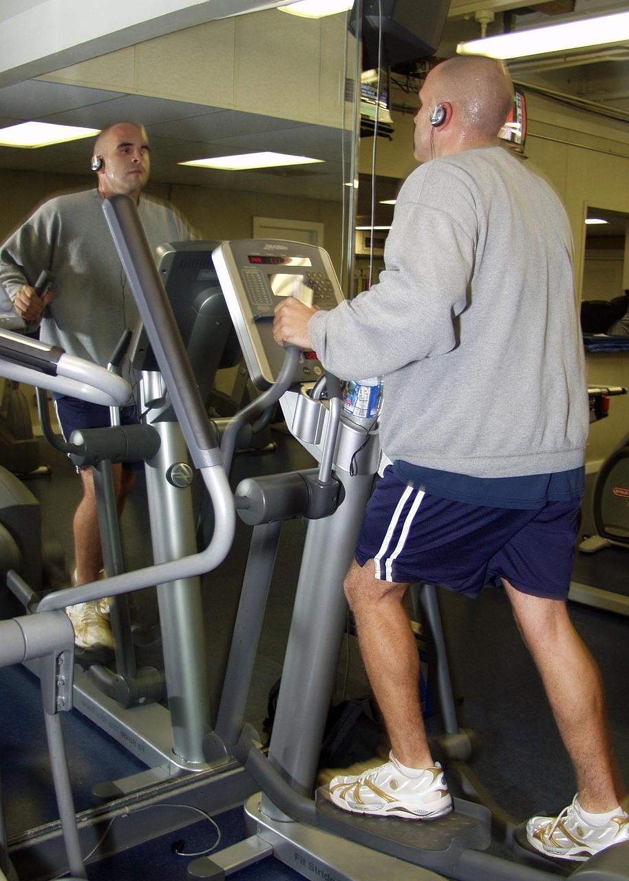 A man running on a treadmill in a gym. He is wearing a grey sweatshirt, blue shorts, and white sneakers. He has a pair of hea...
