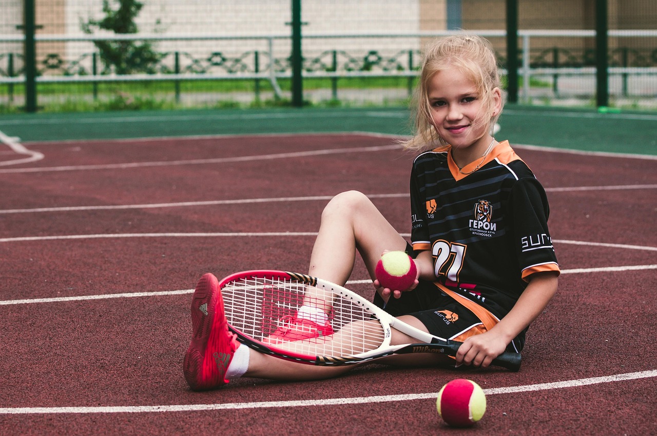 A young girl sitting on a running track with a tennis racket and two tennis balls. She is wearing a black and orange striped ...