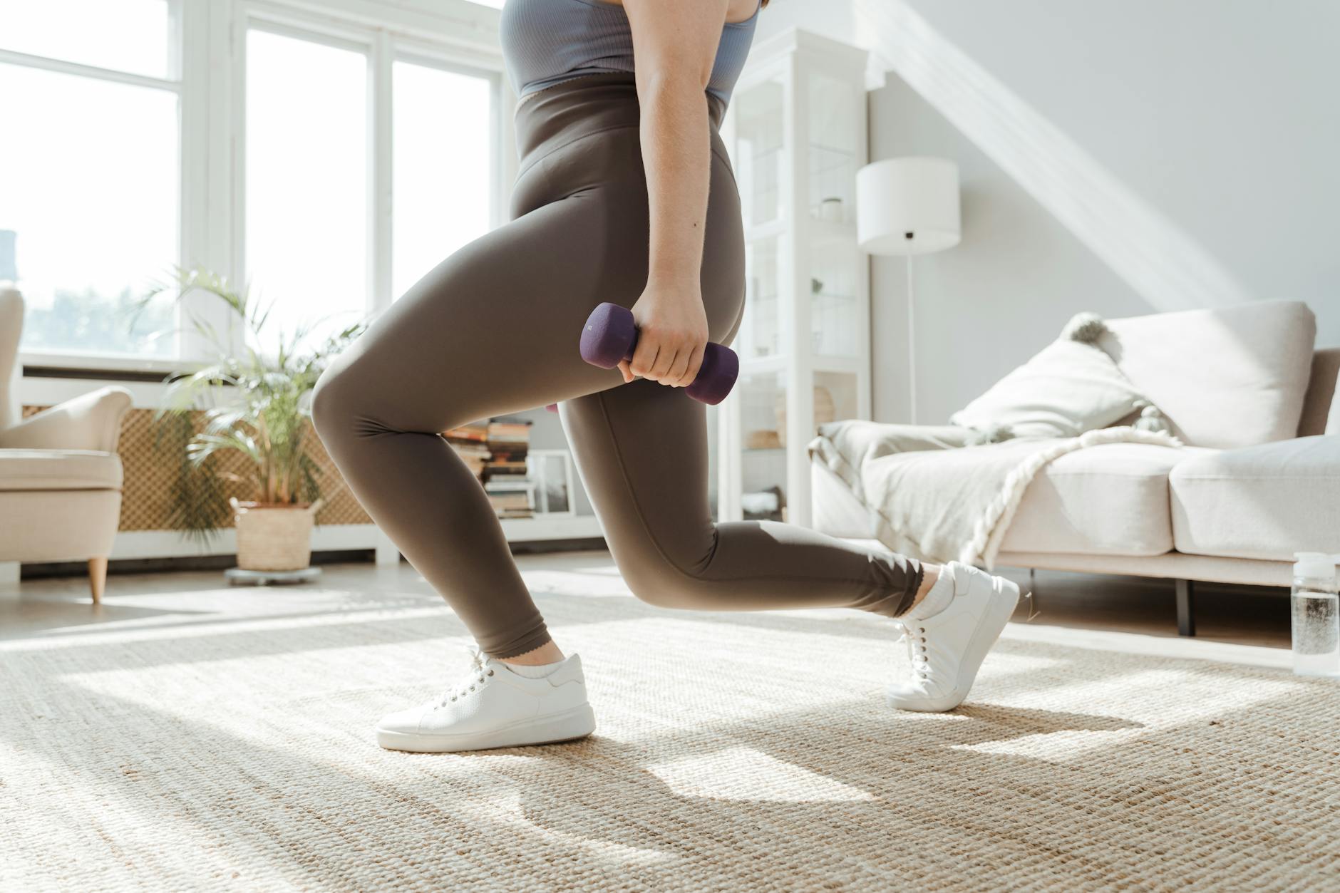 A woman in a living room, performing a lunge exercise with a purple dumbbell. She is wearing a gray tank top and olive green leggings, and white sneakers. The room has a large window on the left side, and a beige sofa on the right side. The floor is covered with a beaded rug, and there is a plant in the corner. The woman is holding the dumbbell in her right hand and her left leg is bent at the knee. She appears to be in the middle of a workout.